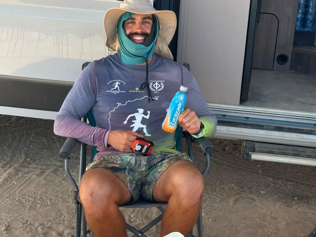 The athlete seated in the shade of the van in a wide-brimmed hat and neck gaiter, holding a drink